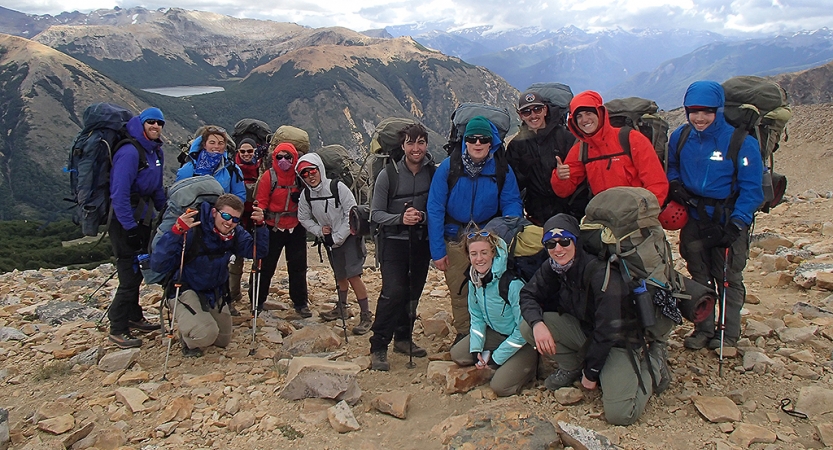 A group of people wearing backpacks pose for a photo while standing on an overlook in front of a vast mountainous landscape.